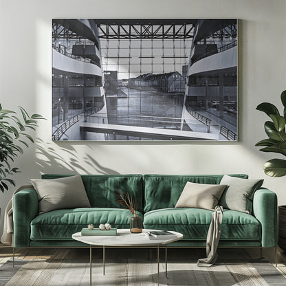 A symmetrical, black and white photograph taken from inside a modern building with curved balconies, looking out through a massive glass wall onto a canal and historic buildings on the opposite bank. A pedestrian bridge crosses the lower part of the frame. Decor
