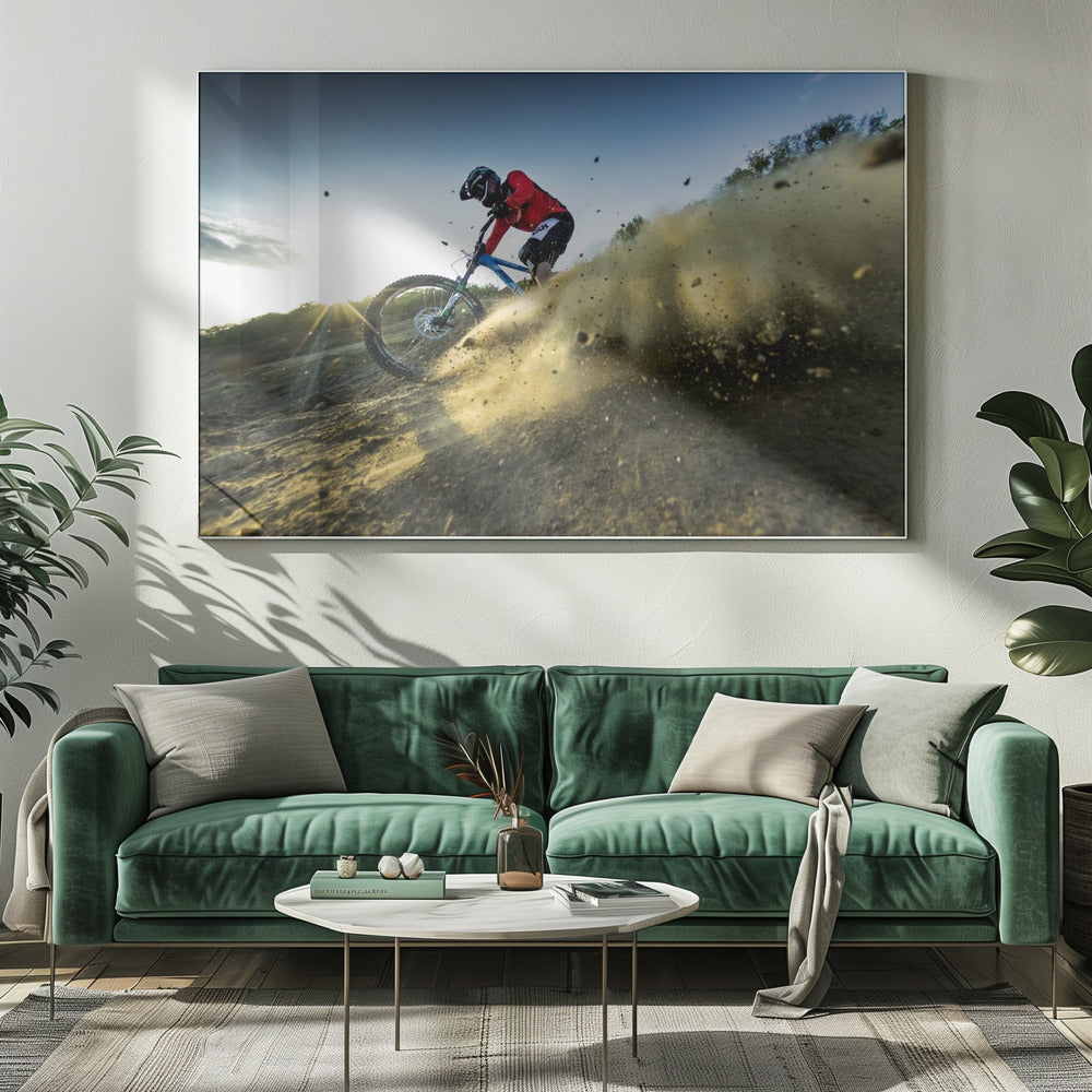 A mountain biker wearing a red jersey and helmet speeds down a steep, dusty trail, kicking up a large cloud of dirt. The sun is setting in the background, creating a lens flare against a clear blue sky. The image is captured from a low angle, emphasizing the action and speed, and is presented within a silver frame. Artwork