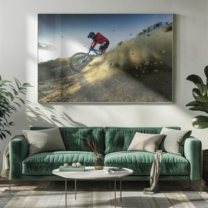A mountain biker wearing a red jersey and helmet speeds down a steep, dusty trail, kicking up a large cloud of dirt. The sun is setting in the background, creating a lens flare against a clear blue sky. The image is captured from a low angle, emphasizing the action and speed, and is presented within a silver frame. Artwork