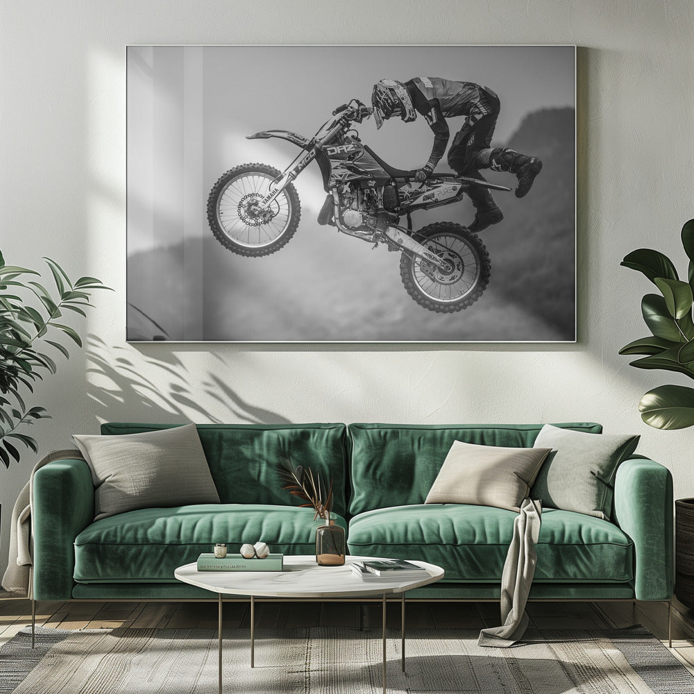 A dynamic black and white photograph of a motocross rider in mid-air, performing a jump. The rider, wearing full protective gear and a helmet, is leaning forward over the handlebars of the dirt bike. The bike is suspended against a blurry, light gray background, emphasizing the action and height of the jump. The image is enclosed in a silver frame. Wall Art