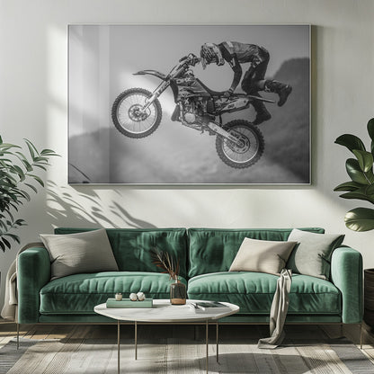 A dynamic black and white photograph of a motocross rider in mid-air, performing a jump. The rider, wearing full protective gear and a helmet, is leaning forward over the handlebars of the dirt bike. The bike is suspended against a blurry, light gray background, emphasizing the action and height of the jump. The image is enclosed in a silver frame. Wall Art