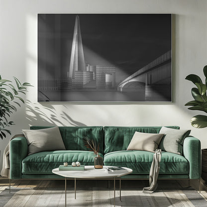 A dramatic black and white photograph of the London skyline featuring The Shard skyscraper and London Bridge. The image is shot at dusk or night, with The Shard brightly lit against a dark, moody sky. The River Thames in the foreground is calm and reflective, captured with a long exposure. Artwork