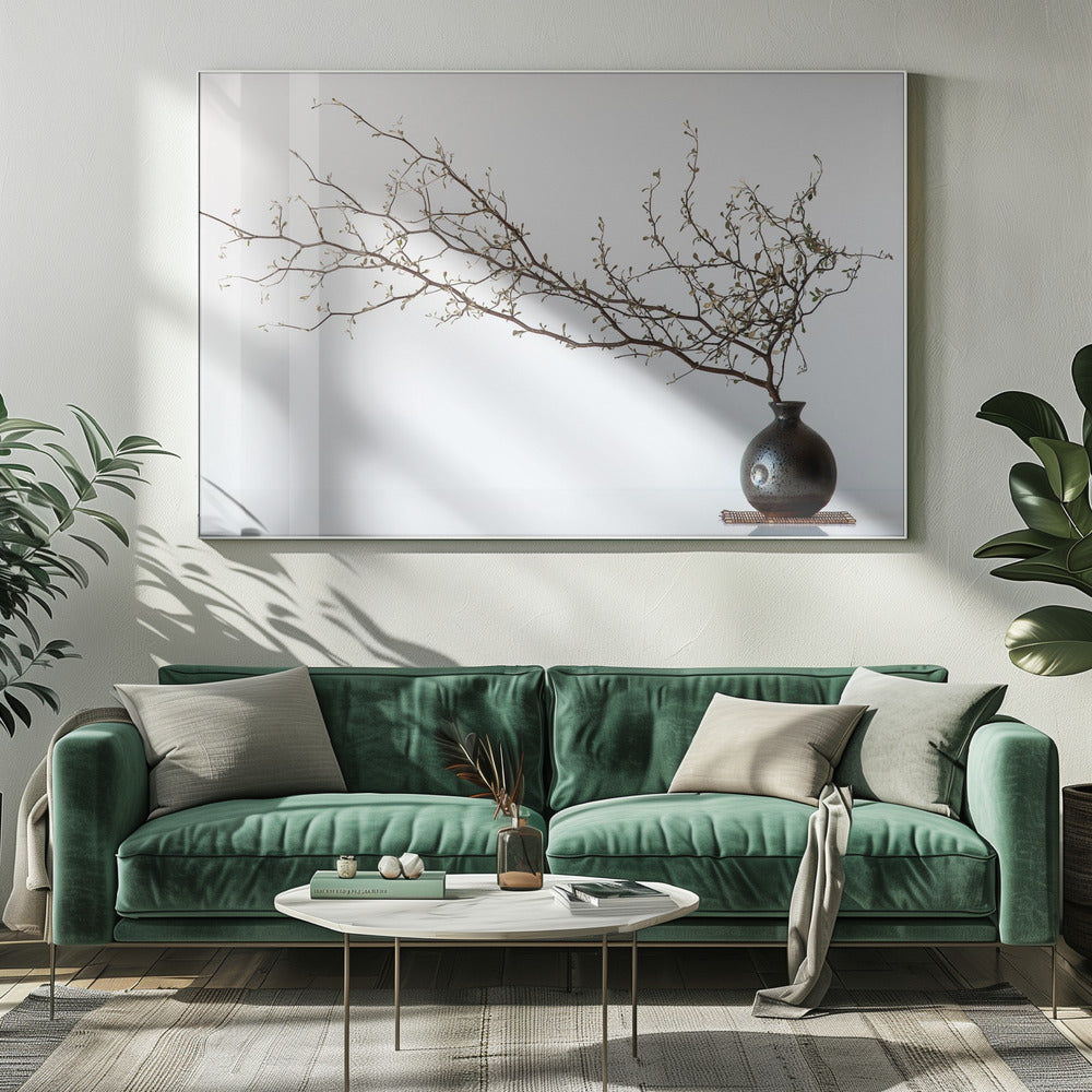 A minimalist still life of a long, delicate branch with small leaves arranged horizontally in a small, dark, round ceramic vase. The background is stark white, creating a high-contrast, serene composition, all within a silver frame. Print
