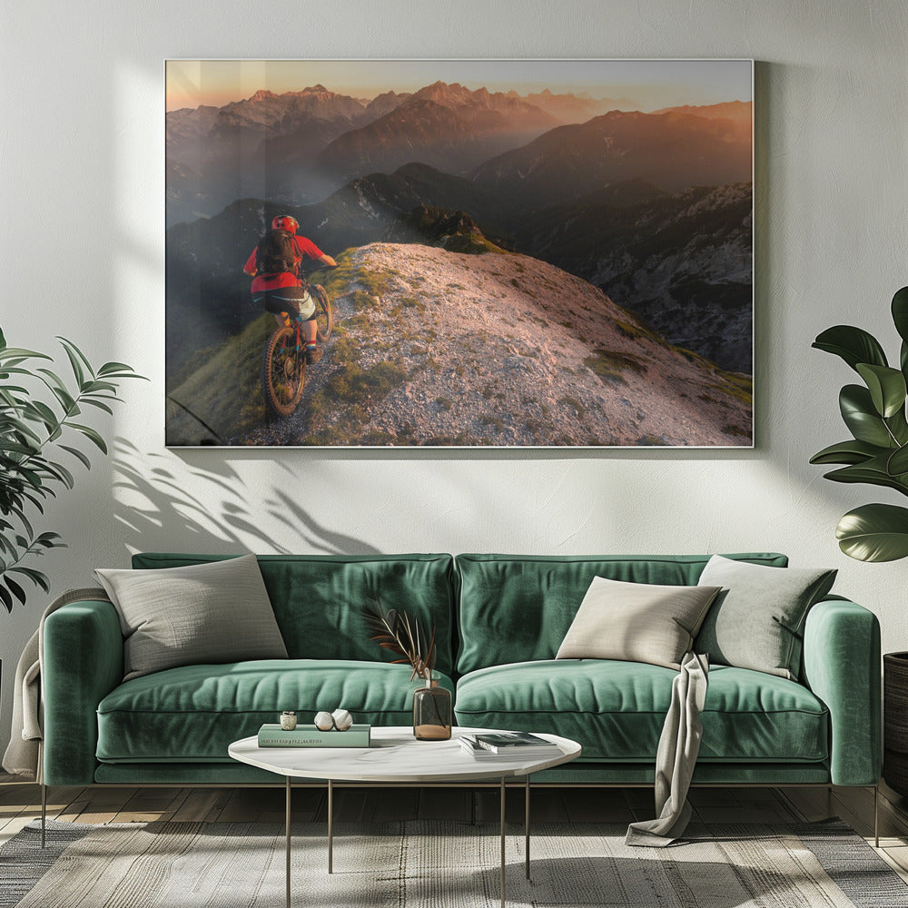 A mountain biker wearing a red shirt and helmet rides along a narrow, rocky mountain ridge at sunset. The background features a vast landscape of layered mountain ranges glowing in the warm, golden light of the setting sun. Poster