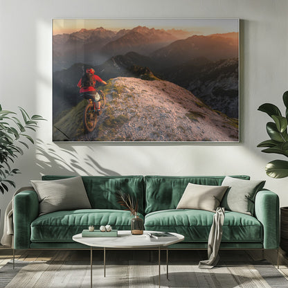 A mountain biker wearing a red shirt and helmet rides along a narrow, rocky mountain ridge at sunset. The background features a vast landscape of layered mountain ranges glowing in the warm, golden light of the setting sun. Poster