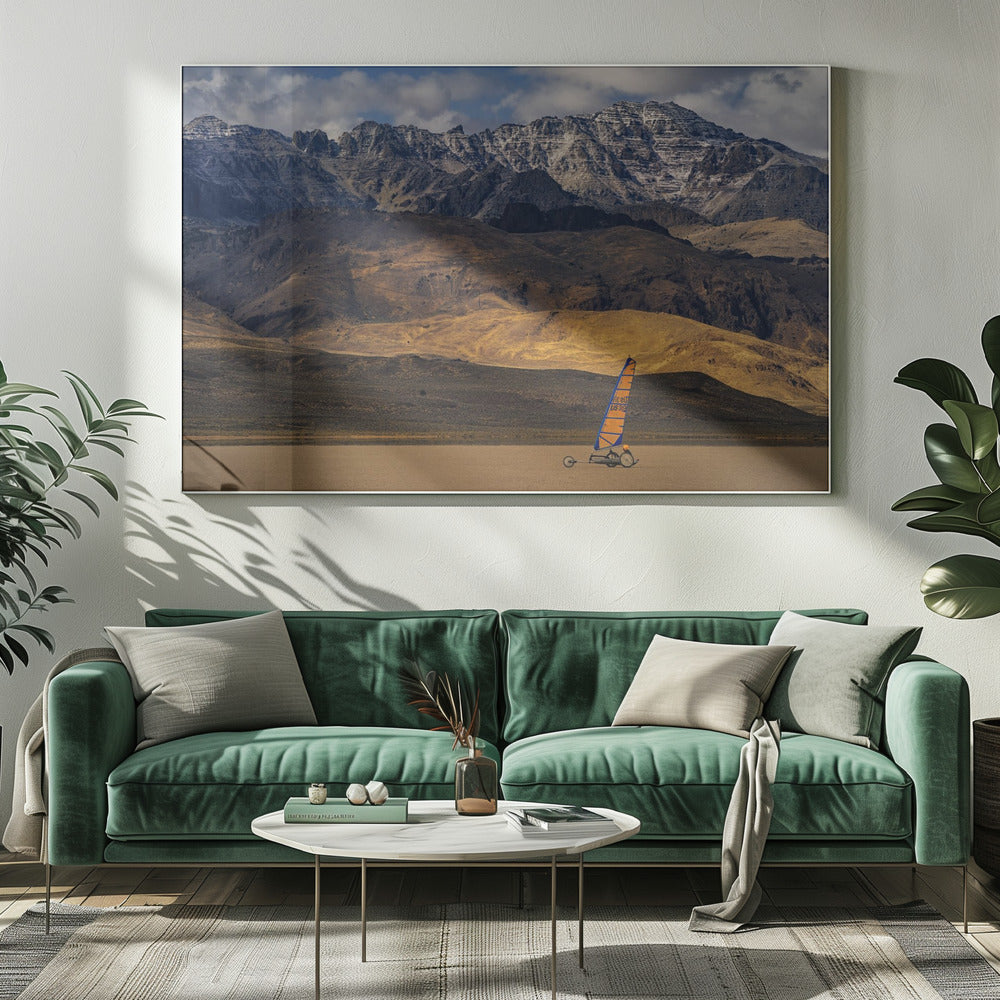 A framed photograph of a land sailor with a bright orange and blue sail on a vast, flat desert floor. In the background, majestic snow-dusted mountains rise against a cloudy blue sky. Artwork