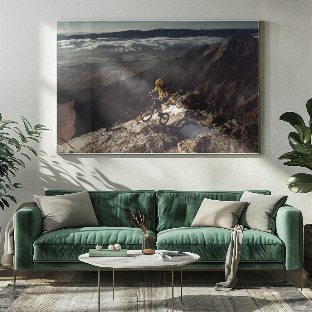 A mountain biker in a yellow helmet and jersey navigates a treacherous, rocky mountain ridge. Below, a vast valley is filled with a sea of clouds, with distant mountain ranges visible on the horizon. Print