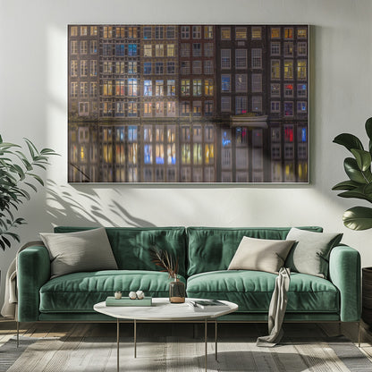 A framed nighttime photograph of a row of traditional Amsterdam canal houses with many illuminated windows, their colorful lights reflecting perfectly in the calm canal water below. A small white boat is moored at the water's edge. Decor