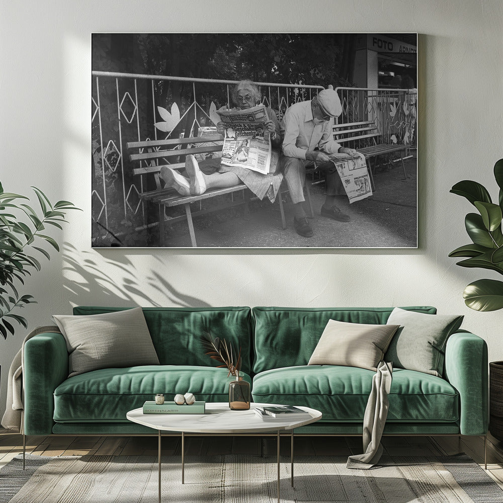 A framed black and white photograph of an elderly couple sitting on a park bench, both deeply absorbed in reading their newspapers. The woman on the left has her feet propped up on the bench, while the man on the right sits next to her, looking down at his paper. Poster