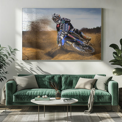 A motocross rider in full gear leans sharply into a turn on a dirt track, kicking up a large spray of mud and dirt from their blue and white Yamaha motorcycle. Artwork