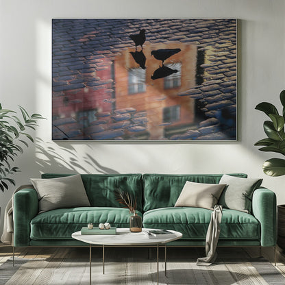 An artistic photograph of two silhouetted birds standing in a puddle on a wet cobblestone street. The puddle reflects an orange building with windows, and the birds' own reflections are visible beneath them, creating ripples in the water. The image is presented in a silver frame. Decor