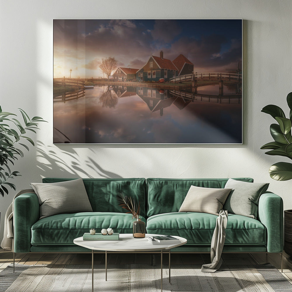 A picturesque landscape of traditional Dutch houses with teal walls and red roofs reflected perfectly in the calm water of a canal at sunset, under a dramatic, cloudy sky. A small wooden bridge crosses the canal, and the scene is enclosed in a silver frame. Artwork