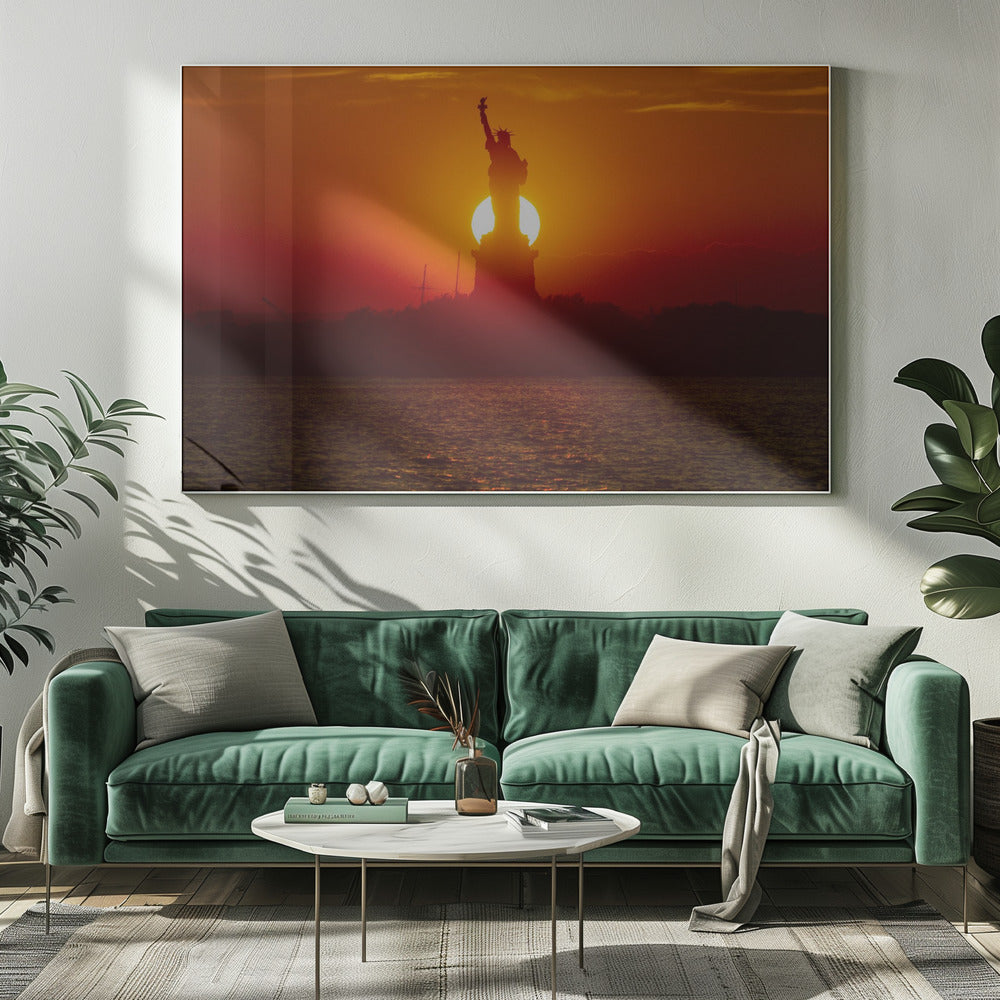 A powerful silhouette of the Statue of Liberty against a fiery orange and red sunset. The sun is perfectly positioned behind the statue's base, creating a brilliant, glowing orb of light. The dark waters in the foreground reflect the vibrant colors of the sky. The photograph is presented within a silver-colored frame. Artwork