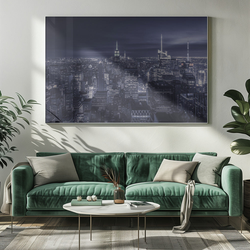 A dramatic nighttime photograph of the New York City skyline, viewed from a high vantage point. The cityscape is a sea of illuminated skyscrapers under a dark, moody sky, with the entire image cast in a cool blue and white monochrome tone and presented in a silver frame. Artwork