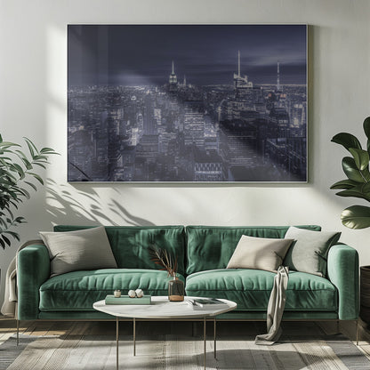 A dramatic nighttime photograph of the New York City skyline, viewed from a high vantage point. The cityscape is a sea of illuminated skyscrapers under a dark, moody sky, with the entire image cast in a cool blue and white monochrome tone and presented in a silver frame. Artwork