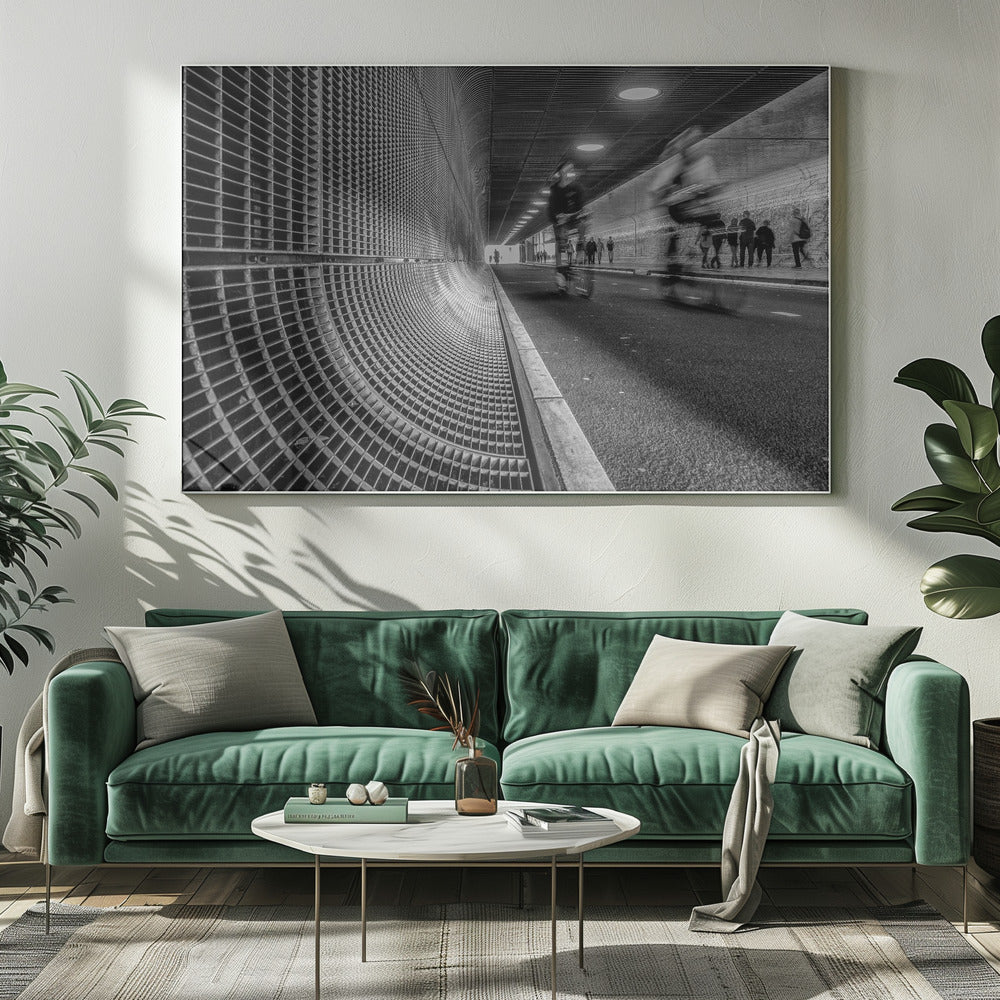 A dynamic black and white photograph from inside a modern tunnel. On the left, a curved metal grate wall creates a strong pattern, while on the right, cyclists are captured in motion blur on a paved path. Pedestrians are visible in the background, walking through the illuminated passage. Poster