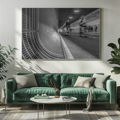 A dynamic black and white photograph from inside a modern tunnel. On the left, a curved metal grate wall creates a strong pattern, while on the right, cyclists are captured in motion blur on a paved path. Pedestrians are visible in the background, walking through the illuminated passage. Poster