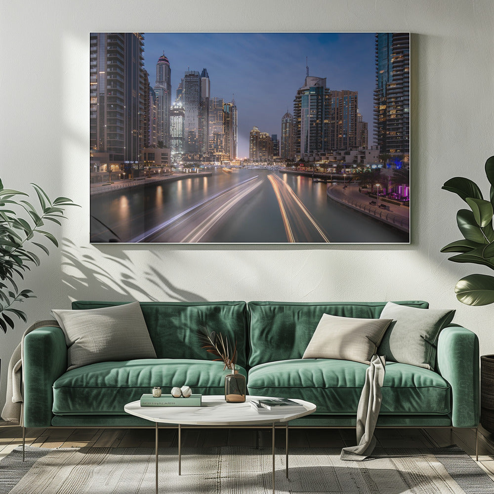 A long-exposure photograph of the Dubai Marina at dusk, framed in silver. Illuminated skyscrapers line a busy waterway, with light trails from moving boats creating long, bright streaks on the water. The sky is a deep twilight blue, and the city's lights cast a warm glow. Decor