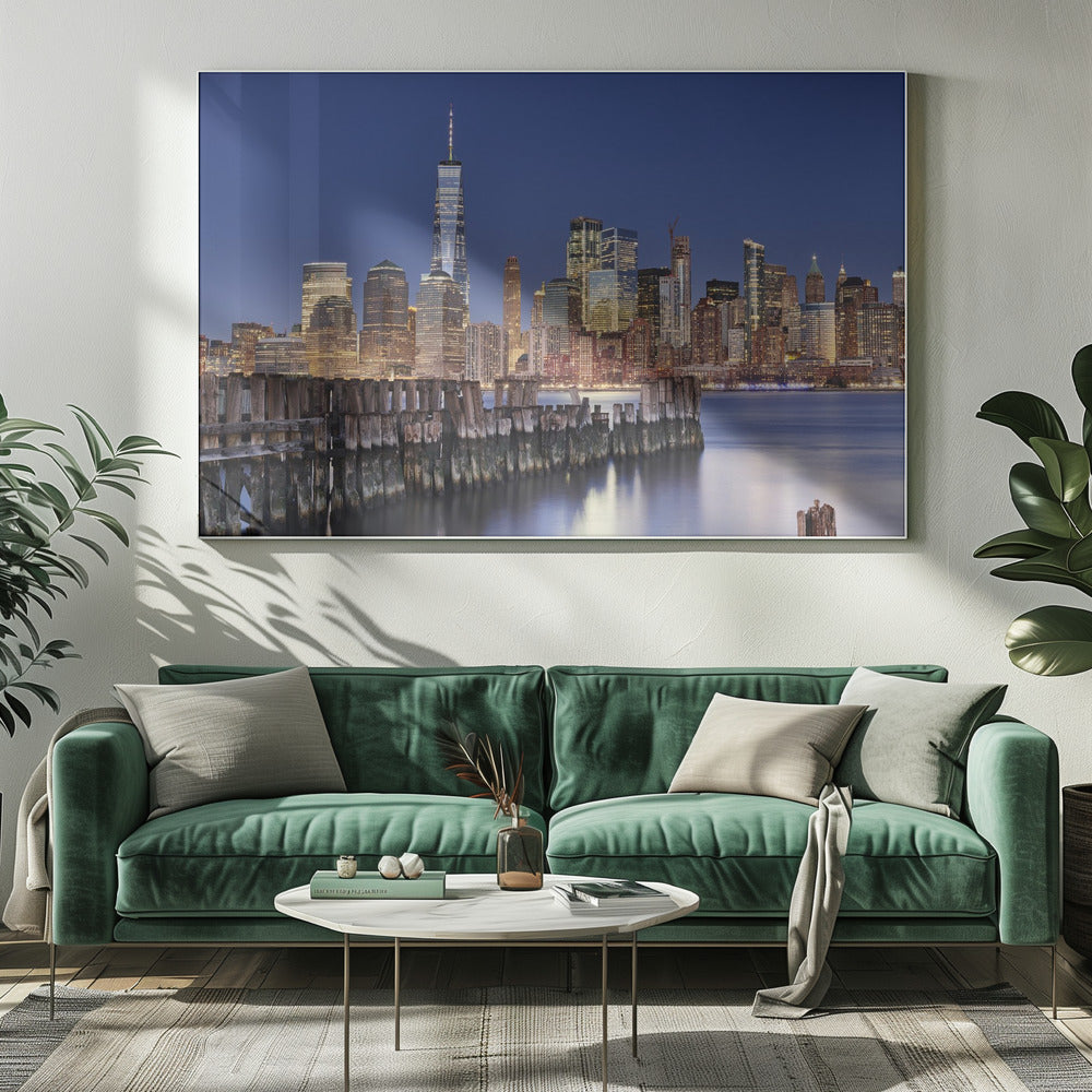 A framed photograph of the Lower Manhattan skyline at dusk. The One World Trade Center and other skyscrapers are brightly lit against a deep blue sky. In the foreground, an old wooden pier extends into the water, which reflects the city lights. Wall Art