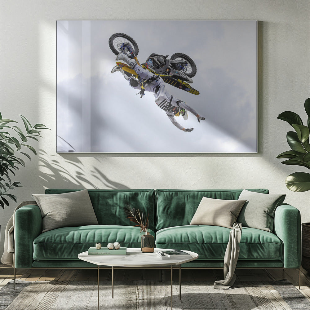 A framed action shot of a freestyle motocross rider in mid-air, performing an upside-down trick against a cloudy sky, with their body hanging below the yellow and black dirt bike. Artwork