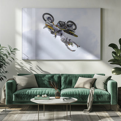 A framed action shot of a freestyle motocross rider in mid-air, performing an upside-down trick against a cloudy sky, with their body hanging below the yellow and black dirt bike. Artwork
