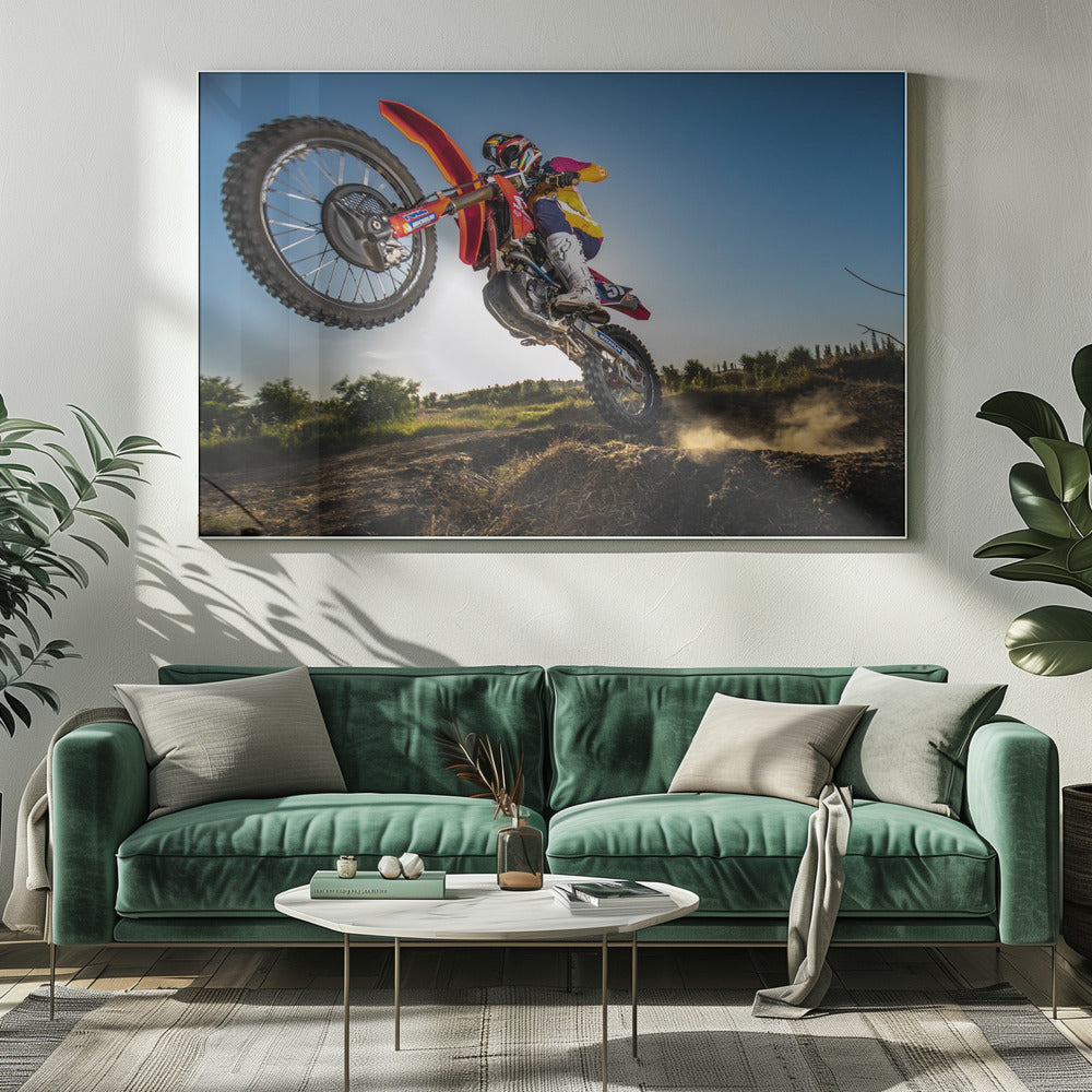 A dynamic, low-angle shot of a motocross rider in mid-air, jumping over a dirt mound. The rider is on a red, white, and blue dirt bike, wearing colorful racing gear and a helmet. The bike's front wheel is high in the air against a clear blue sky, while the back wheel is just clearing the jump, kicking up a cloud of dust. The background features a green, rural landscape. Wall Art