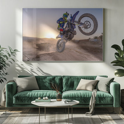A motocross rider in blue and yellow gear is captured mid-air on a blue dirt bike, performing a jump against a dusty landscape with a setting sun in the background. The image is enclosed in a silver frame. Wall Art