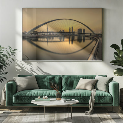 A sweeping view of the Dubai Water Canal Bridge at sunset, its white arch and cables stretching over the calm water. The city skyline, including the Burj Khalifa, is silhouetted against the golden sky, with the entire scene creating a perfect reflection in the water below. Print