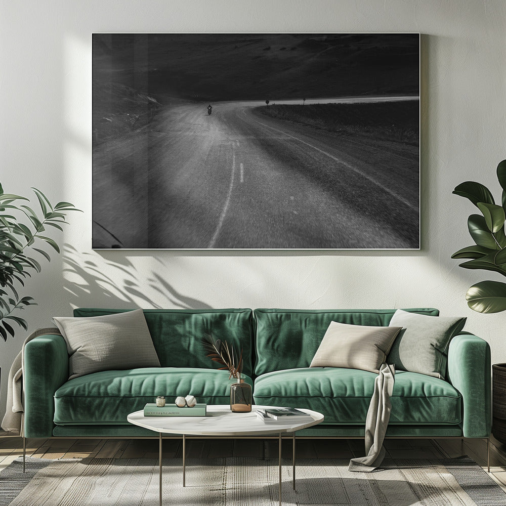 A moody, black and white photograph capturing a lone motorcyclist from behind as they navigate a curving road through a dark, hilly terrain, framed in silver. Decor