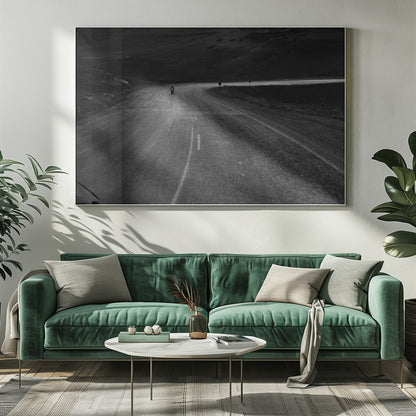 A moody, black and white photograph capturing a lone motorcyclist from behind as they navigate a curving road through a dark, hilly terrain, framed in silver. Decor