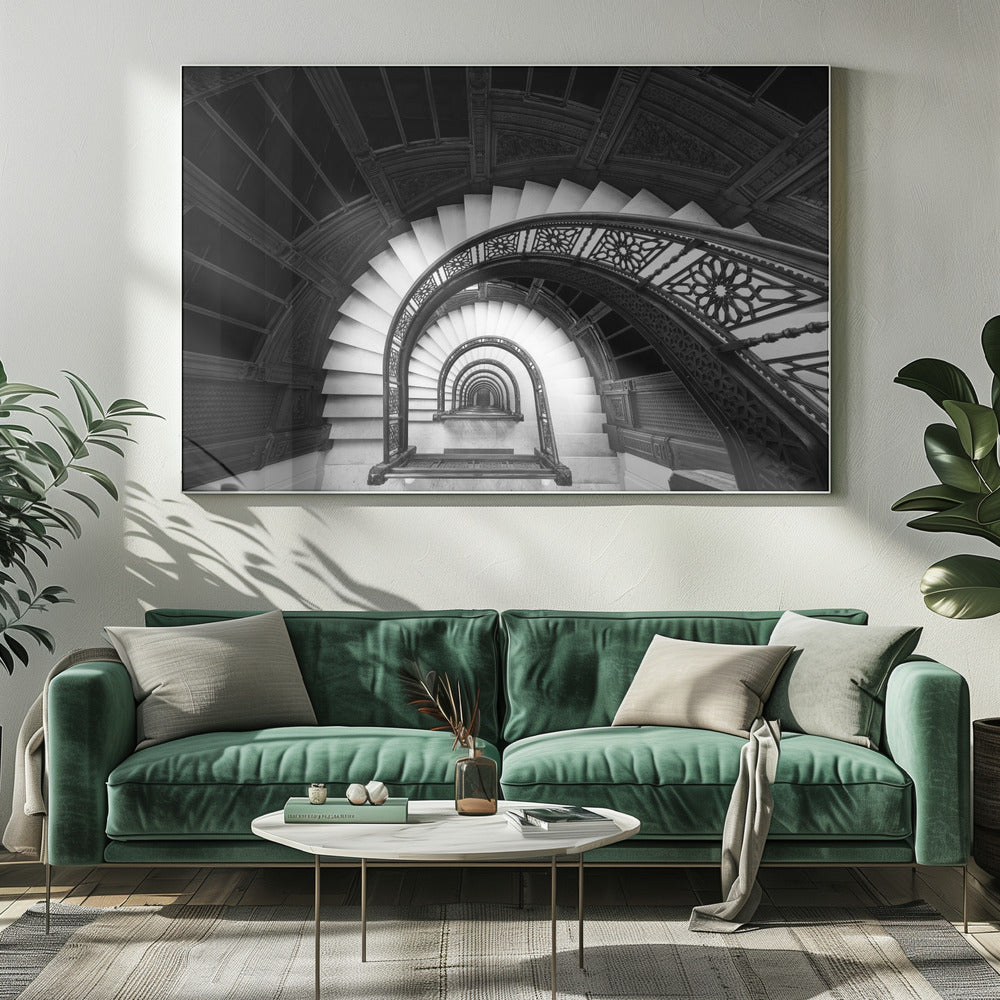 A mesmerizing black and white photograph shot from a high angle looking down into the center of a spiral staircase. The bright white marble steps contrast with the dark, ornate metal railing, creating a repeating arched pattern that draws the eye downward into a seemingly endless tunnel. The image is presented within a silver-colored frame. Artwork