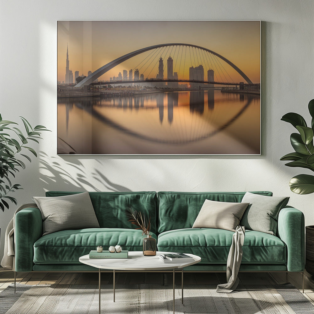 A panoramic photograph of the Dubai skyline at sunrise, viewed from across the water. A large, modern arched bridge spans the canal in the foreground, and its reflection creates a perfect oval shape in the calm water. In the background, skyscrapers, including the Burj Khalifa, are silhouetted against a glowing orange and yellow sky. The entire scene is presented within a silver picture frame. Wall Art