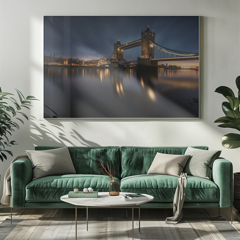 A long-exposure photograph of London's Tower Bridge at dusk, brilliantly illuminated with golden lights that cast shimmering reflections on the calm River Thames. The sky is a deep, dark blue, and the city skyline glows in the background, all enclosed within a silver frame. Print