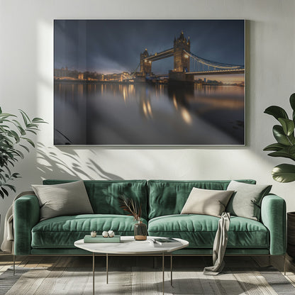 A long-exposure photograph of London's Tower Bridge at dusk, brilliantly illuminated with golden lights that cast shimmering reflections on the calm River Thames. The sky is a deep, dark blue, and the city skyline glows in the background, all enclosed within a silver frame. Print
