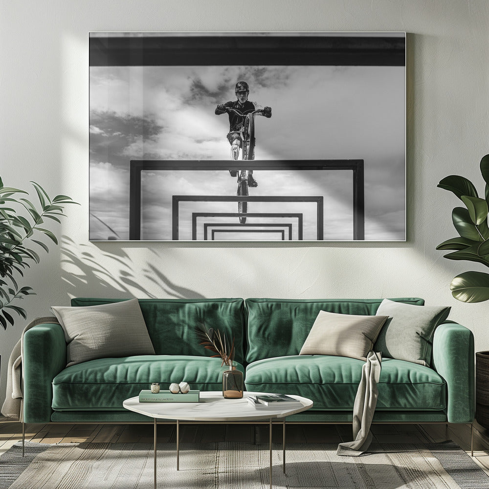 A striking black and white photograph taken from a low angle, showing a cyclist in full gear balancing on a mountain bike. The cyclist is positioned centrally above a series of black rectangular frames that create a receding, tunnel-like visual effect against a cloudy sky. The entire image is presented within a silver picture frame. Artwork
