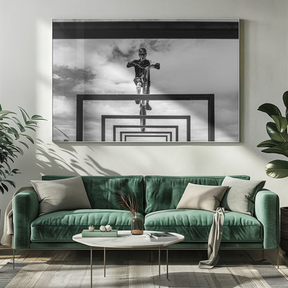 A striking black and white photograph taken from a low angle, showing a cyclist in full gear balancing on a mountain bike. The cyclist is positioned centrally above a series of black rectangular frames that create a receding, tunnel-like visual effect against a cloudy sky. The entire image is presented within a silver picture frame. Artwork