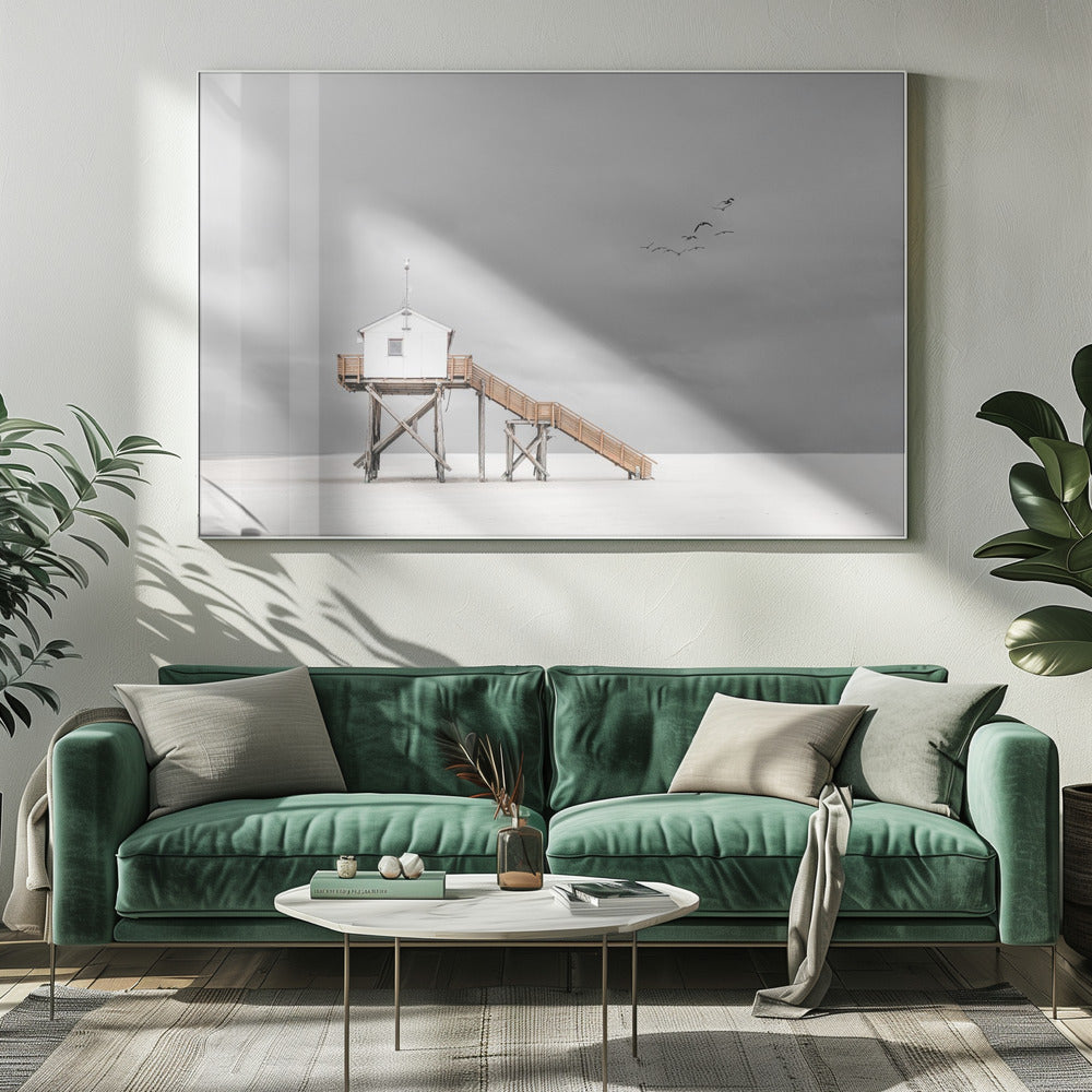 A minimalist, desaturated photograph of a small white cabin on tall wooden stilts on a vast, empty beach. A long wooden staircase leads up to the cabin. The sky is a flat, overcast grey, and a flock of dark birds is visible flying in the distance. Print
