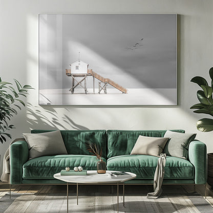 A minimalist, desaturated photograph of a small white cabin on tall wooden stilts on a vast, empty beach. A long wooden staircase leads up to the cabin. The sky is a flat, overcast grey, and a flock of dark birds is visible flying in the distance. Print