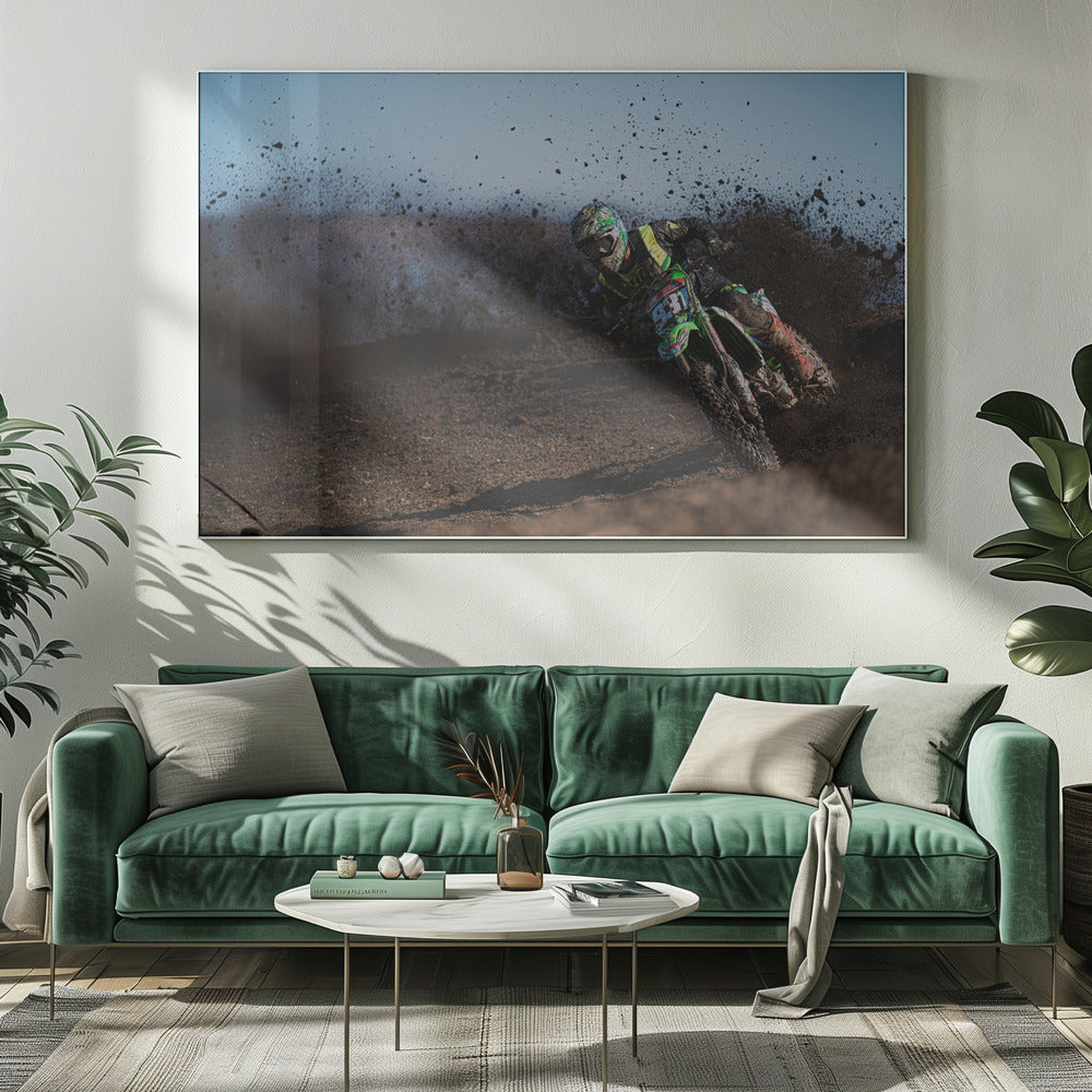 A motocross rider in green and black gear leans hard into a turn on a dirt track, kicking up a large spray of mud against a clear blue sky. Decor