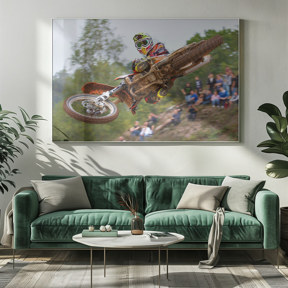 A dynamic action shot of a motocross racer in mid-air, leaning into a turn with their muddy orange dirt bike. The rider wears a colorful helmet and racing gear, with a blurred background of green trees and spectators watching from a dirt hill. The photo is enclosed in a silver frame. Decor