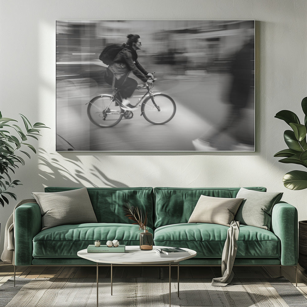 A dynamic black and white photograph capturing a person in profile as they cycle swiftly through a city. The background is a motion blur of urban scenery, highlighting the cyclist's speed. The individual wears a leather jacket, sunglasses, a backpack, and has their hair tied up. The artwork is enclosed in a silver frame. Wall Art
