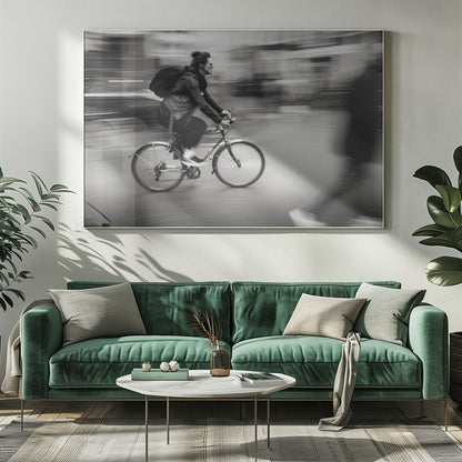 A dynamic black and white photograph capturing a person in profile as they cycle swiftly through a city. The background is a motion blur of urban scenery, highlighting the cyclist's speed. The individual wears a leather jacket, sunglasses, a backpack, and has their hair tied up. The artwork is enclosed in a silver frame. Wall Art