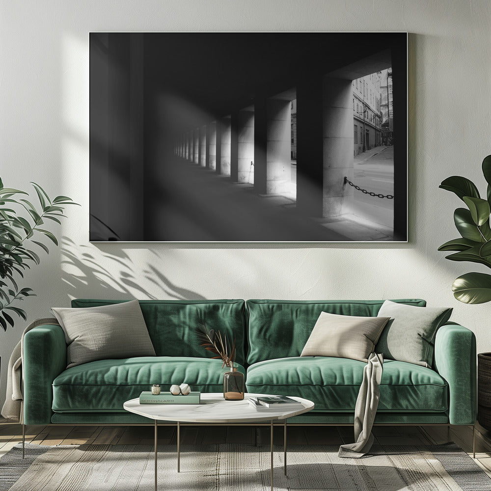 A high-contrast, black and white photograph from the perspective of a dark, covered walkway lined with a row of thick pillars. Light from an adjacent street highlights the pillars, creating a dramatic play of light and shadow and a strong sense of depth. Print