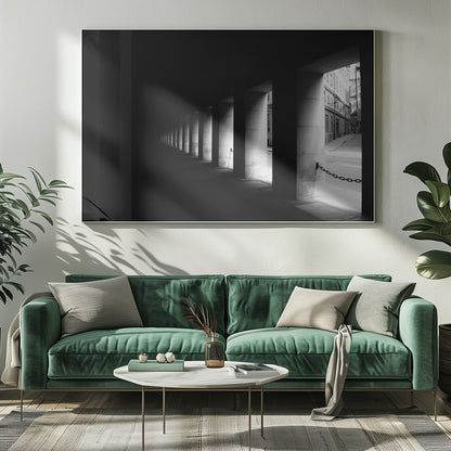 A high-contrast, black and white photograph from the perspective of a dark, covered walkway lined with a row of thick pillars. Light from an adjacent street highlights the pillars, creating a dramatic play of light and shadow and a strong sense of depth. Print