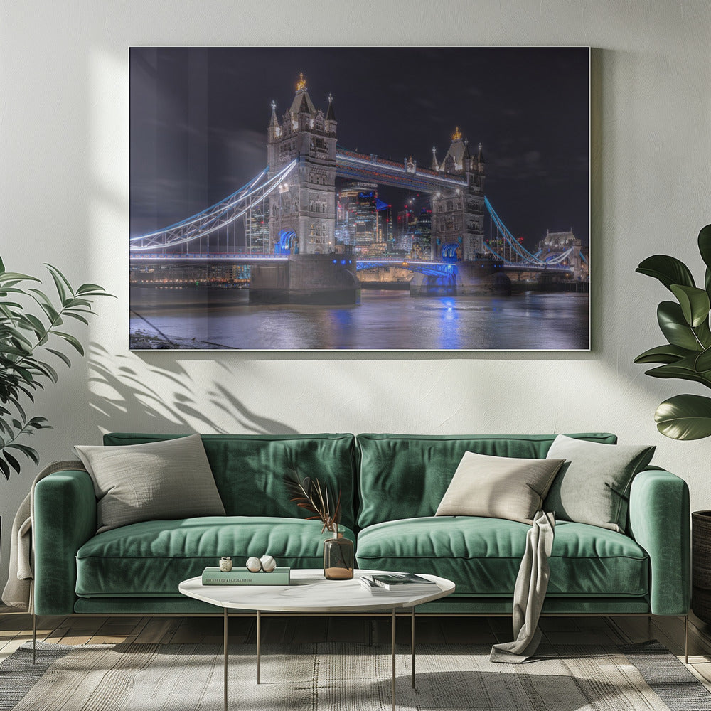 A vibrant night photograph of the iconic Tower Bridge in London, illuminated with bright white and electric blue lights. The dark River Thames in the foreground reflects the bridge and the lit-up modern city skyline in the background, all under a dark, cloudy sky. The image is enclosed in a silver frame. Decor