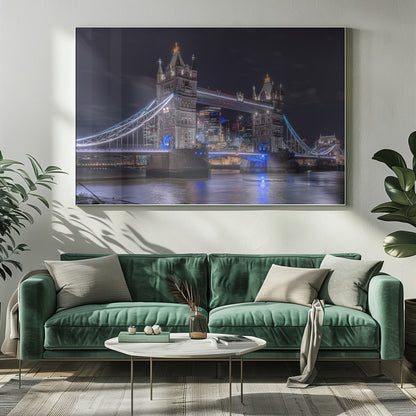 A vibrant night photograph of the iconic Tower Bridge in London, illuminated with bright white and electric blue lights. The dark River Thames in the foreground reflects the bridge and the lit-up modern city skyline in the background, all under a dark, cloudy sky. The image is enclosed in a silver frame. Decor