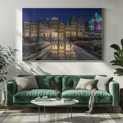 A vibrant night-time photograph of a canal in Amsterdam, flanked by two tour boats in the foreground. Traditional Dutch canal houses are brightly lit, casting warm yellow reflections on the dark water under a deep blue sky. Print