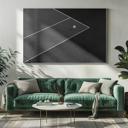 A minimalist black and white overhead photograph of a white ball resting in the corner of a dark tennis or paddle court. The composition focuses on the strong geometric angles created by the white boundary lines against the dark, textured asphalt, all within a silver frame. Artwork