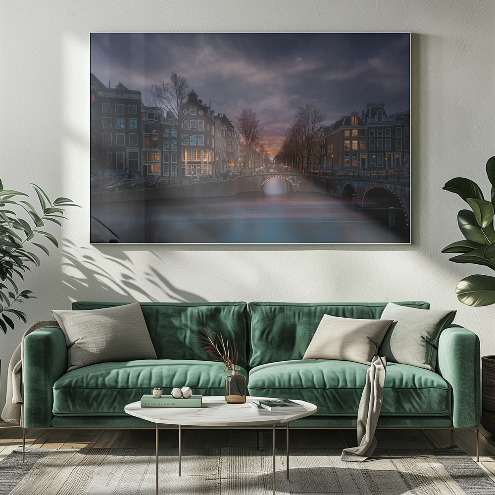 A dramatic long-exposure photograph of an Amsterdam canal at twilight. An arched bridge spans the water, lined by historic Dutch buildings with glowing windows. The sky is filled with dark, moody clouds above a fading orange and purple sunset, and red light trails from boats are blurred on the water's surface. Poster