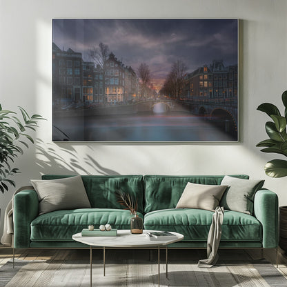 A dramatic long-exposure photograph of an Amsterdam canal at twilight. An arched bridge spans the water, lined by historic Dutch buildings with glowing windows. The sky is filled with dark, moody clouds above a fading orange and purple sunset, and red light trails from boats are blurred on the water's surface. Poster